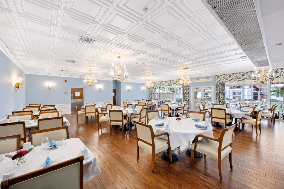 A bright and spacious dining room with multiple tables covered in white tablecloths, each set with glasses, cups, and blue napkins. The room features wooden chairs with cream cushions, elegant chandeliers hanging from a white coffered ceiling, light blue walls, and large windows with floral curtains allowing natural light to fill the space.