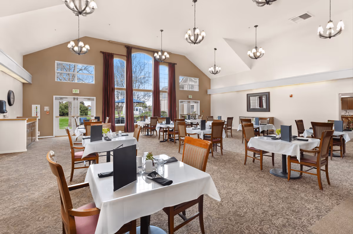 A spacious dining room with multiple tables covered in white tablecloths, each set with black menus, napkins, and glassware. The room features large windows with red curtains, allowing natural light to brighten the space. Several chandeliers hang from the high ceiling, and the carpet has a patterned design. There are wooden chairs around each table, and the room has a warm, inviting atmosphere.