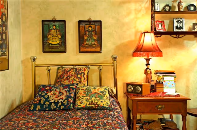 A cozy bedroom with a brass bed frame adorned with colorful patterned pillows and a matching bedspread. Above the bed are two framed artworks depicting traditional figures. To the right of the bed is a wooden nightstand with a vintage radio, a stack of books, and a table lamp with a red fringed shade. A wooden shelf above the nightstand holds framed photos and decorative items.