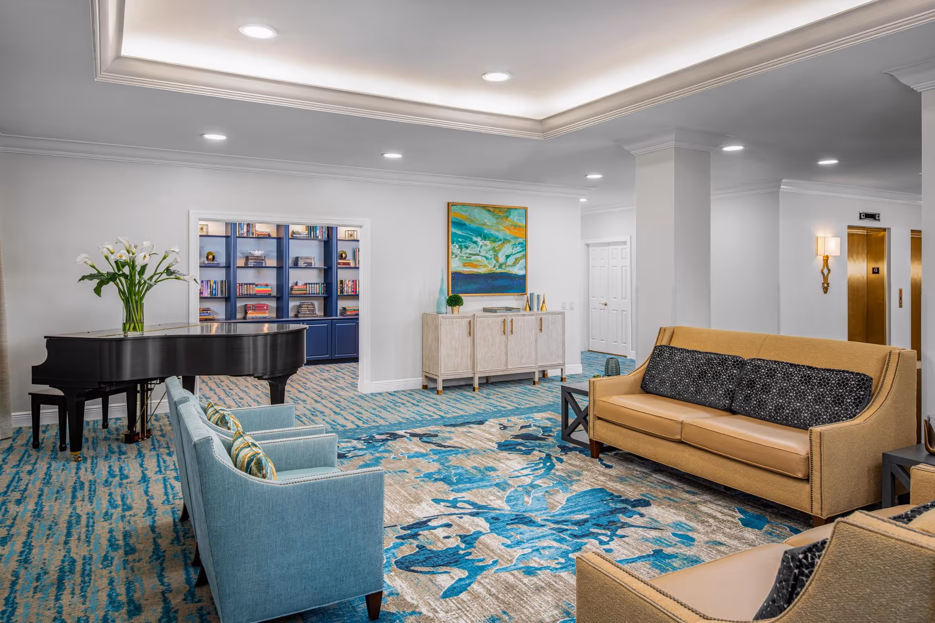 A bright and spacious living room area with a blue and beige patterned carpet, a black grand piano with a vase of white flowers on top, two light blue armchairs with patterned cushions, a tan leather sofa with black patterned cushions, and a beige sideboard with decorative items and a colorful abstract painting above it. In the background, there is a doorway leading to a room with blue bookshelves filled with books and decorative objects. The walls are white, and there are recessed ceiling lights and wall sconces near two elevator doors.