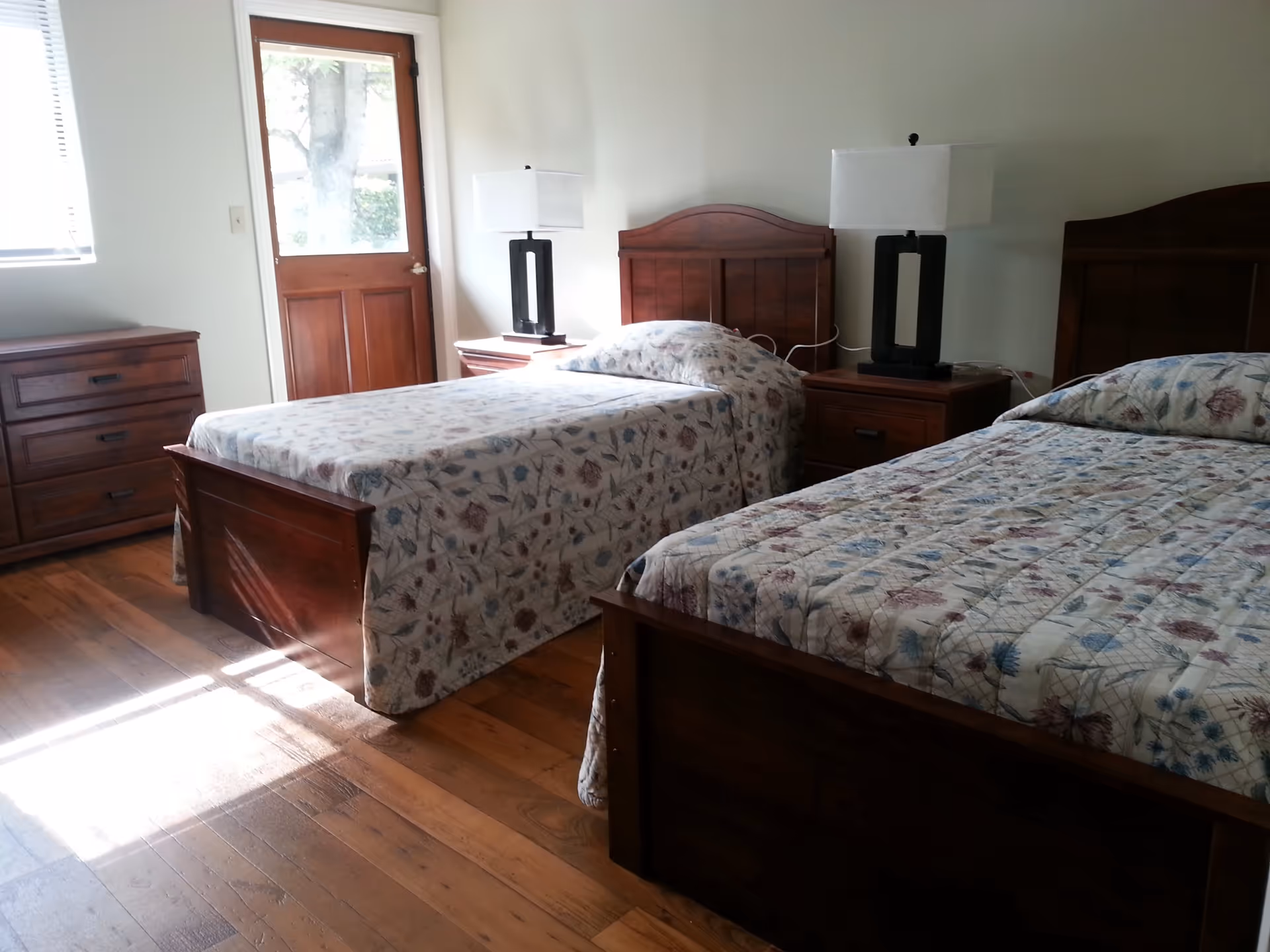 A bedroom with two single beds featuring wooden headboards and floral bedspreads. Between the beds is a wooden nightstand with a modern black lamp with a white shade. To the left, there is a wooden dresser and a door with a window letting in natural light. The floor is wooden and the walls are painted white.