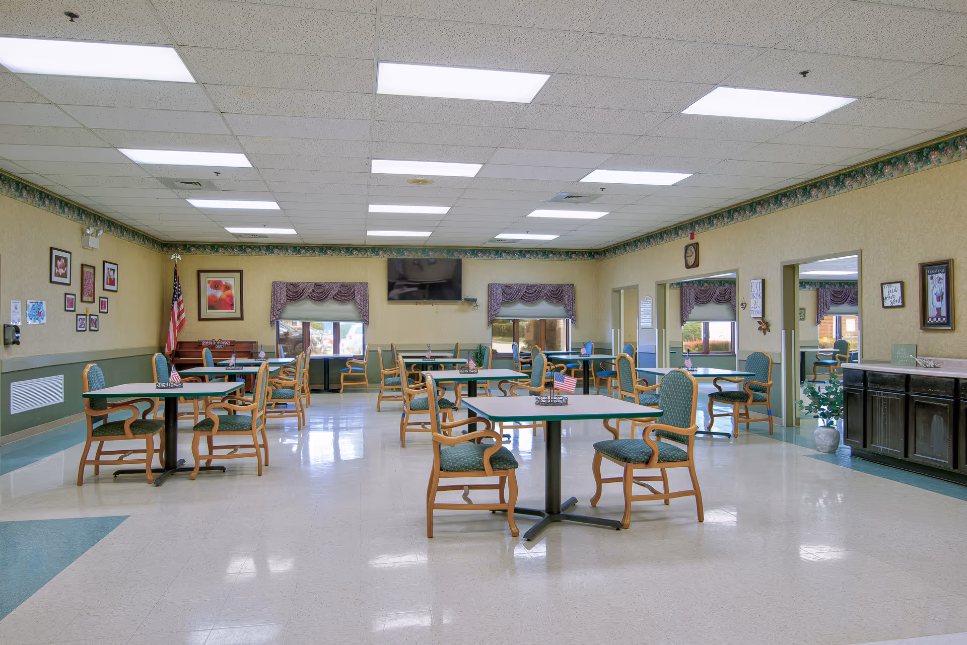Bright communal dining room with multiple square tables and green-upholstered chairs, a wall-mounted TV, and an American flag.