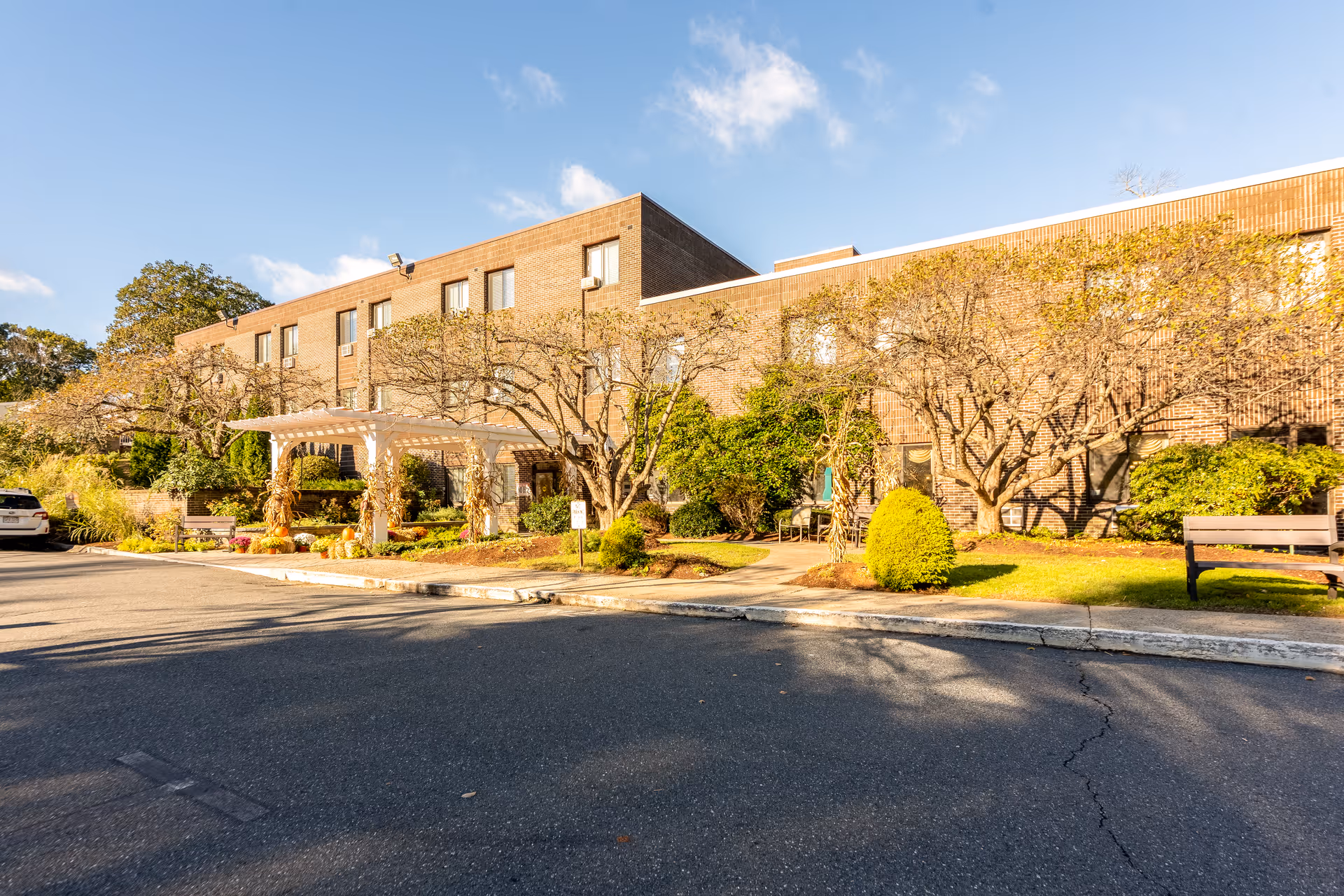 Exterior view of a two-story brick building with a white pergola at the entrance, surrounded by landscaped greenery and trees. There is a paved driveway and a parked white car on the left side. The sky is clear with a few clouds.