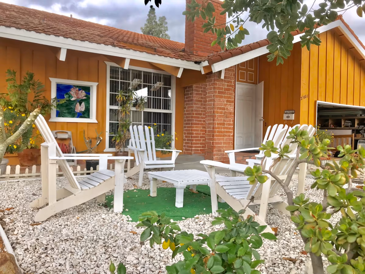 Outdoor seating area with four white wooden chairs arranged around a small white table on a green mat, surrounded by white gravel. The background shows a yellow house with a red tiled roof, a brick chimney, a white door, and a window with bars. There are plants and greenery around the seating area.