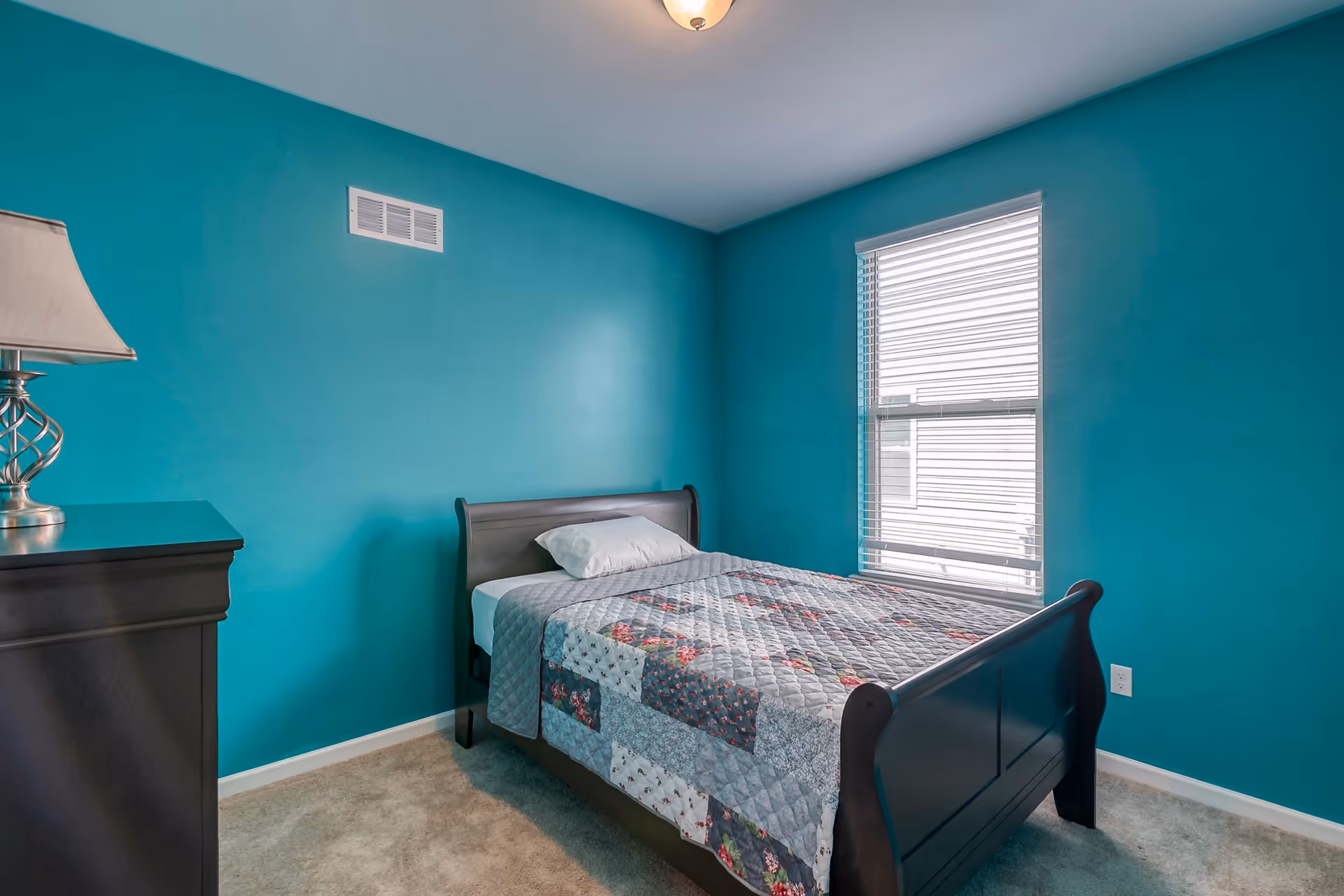 A small bedroom with teal walls, a single dark wooden bed with a patchwork quilt and a white pillow, a window with white blinds letting in natural light, a beige carpeted floor, and a dark wooden dresser with a silver lamp on top.