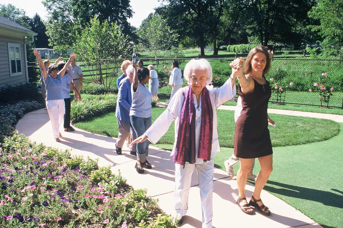 A group of elderly people and a younger woman are walking and holding hands on a curved outdoor pathway surrounded by green grass, flowers, and trees on a sunny day.