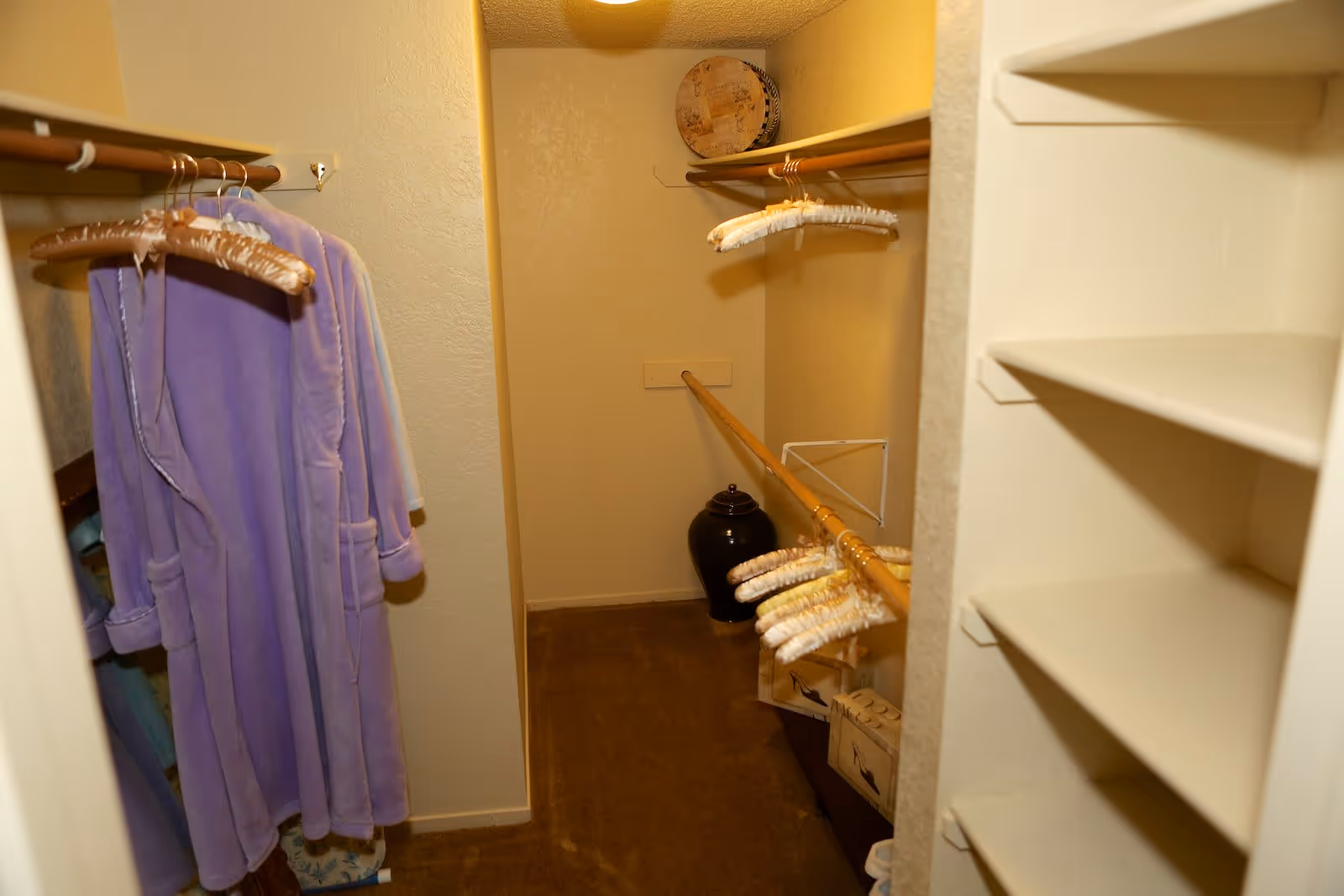 A small walk-in closet with beige walls and carpeted floor. There are wooden rods on both sides with several padded hangers and a purple robe hanging on the left side. On the right, there are empty shelves. At the back of the closet, there is a black decorative vase and some boxes on the floor.