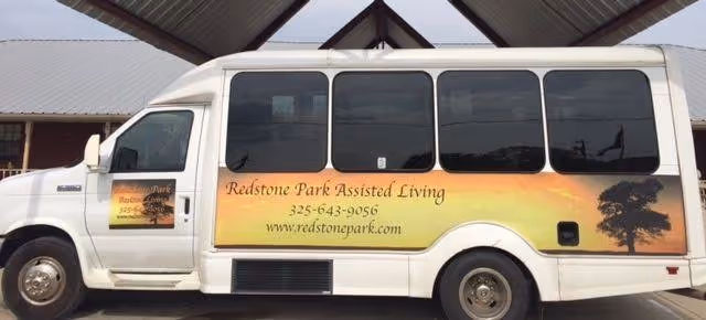 White shuttle van for Redstone Park Assisted Living parked under a metal canopy, showing the facility name, phone number, and website on its side.