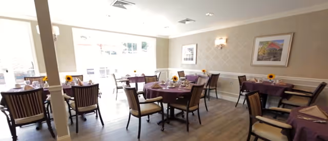 A bright dining room with several round tables covered with purple tablecloths, each set with plates, glasses, and napkins. Each table has a small vase with a sunflower. The room has light-colored walls with a framed picture and wall sconces, and large windows letting in natural light.