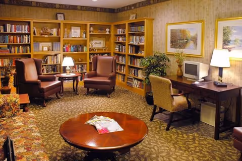A cozy living room area with two brown leather armchairs and a floral patterned sofa around a round wooden coffee table with magazines on it. There are built-in wooden bookshelves filled with books and decorative items along the back wall. A wooden desk with a chair, a small computer, and a lamp is positioned against the right wall, which is decorated with framed landscape paintings. The room is warmly lit with table lamps and has a patterned carpet.