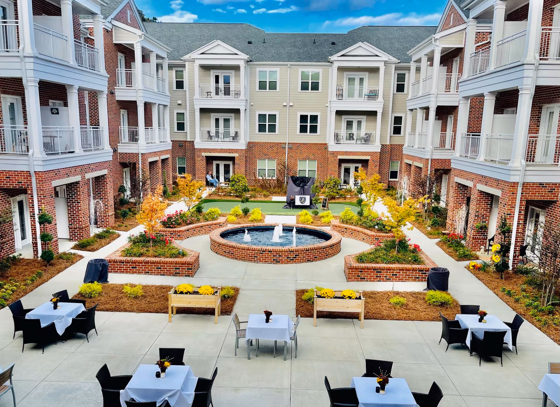 Central landscaped courtyard with a circular fountain, outdoor tables with white tablecloths, and surrounding multi-story brick senior living building with balconies.