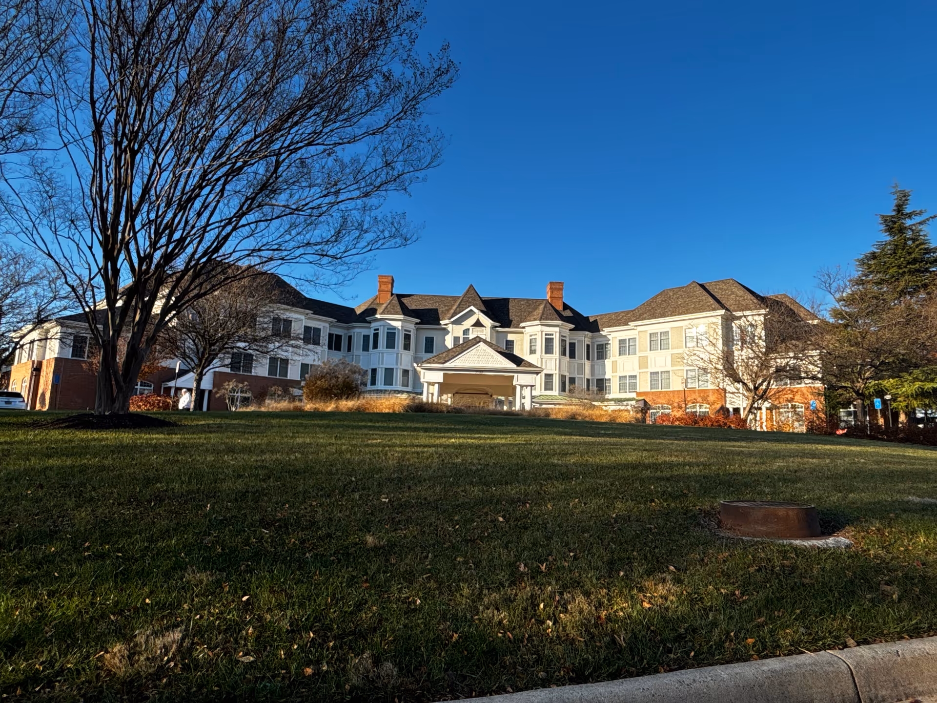 Multi-story white building with a covered entrance, a wide grassy lawn and leafless trees under a clear blue sky.