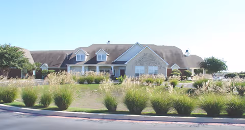 Exterior view of a single-story building with a sloped roof and multiple dormer windows, surrounded by landscaped greenery and ornamental grasses in front.