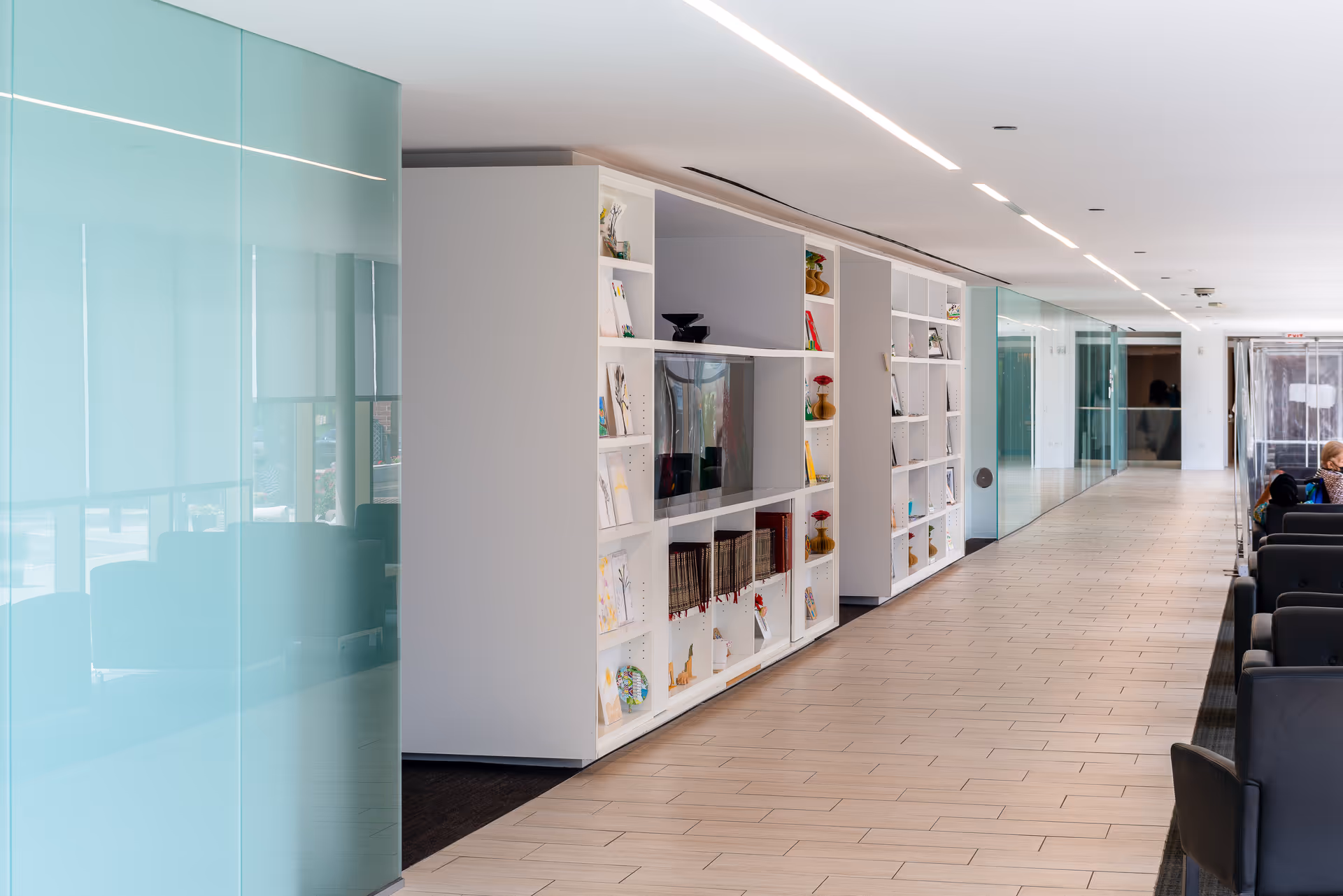 Bright modern interior corridor with built-in white shelving, glass walls, and rows of chairs.