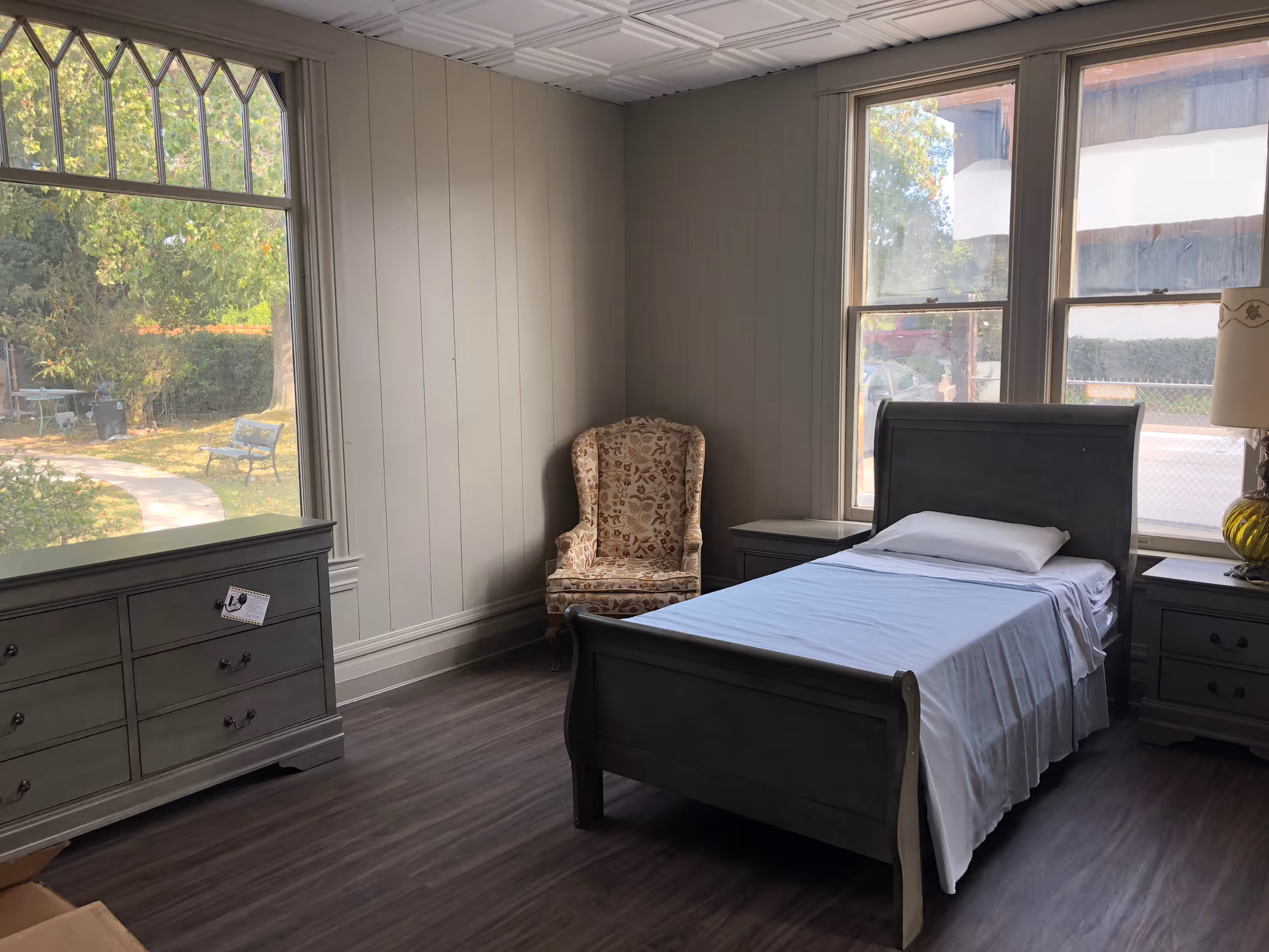 A simple bedroom with a single bed made with white sheets and a pillow, two matching gray nightstands on either side of the bed, a gray dresser with drawers, and a floral upholstered armchair in the corner. Large windows allow natural light to fill the room, showing a view of a garden path and outdoor benches.