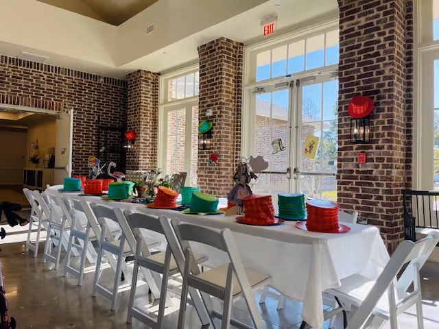 A long dining table set with a white tablecloth and decorated with colorful hats and themed centerpieces in a bright room with large windows and brick walls. White folding chairs are arranged along both sides of the table, and sunlight streams through the windows.