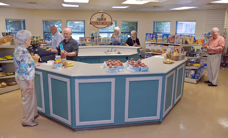 Interior of a small store area named 'Village Store' inside a senior living facility. Several elderly people are shopping and interacting near a central counter displaying fresh fruit and groceries. Shelves stocked with various food items and snacks are visible in the background.