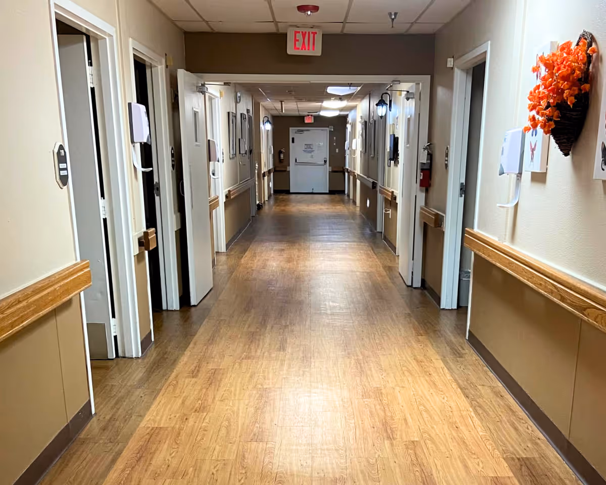 A long, clean hallway in a healthcare or rehabilitation facility with wooden flooring and beige walls. Several open doors line both sides of the hallway, and handrails run along the walls. There are wall-mounted dispensers and decorations, including a basket with orange flowers on the right wall. An exit sign is visible at the far end above a closed door.