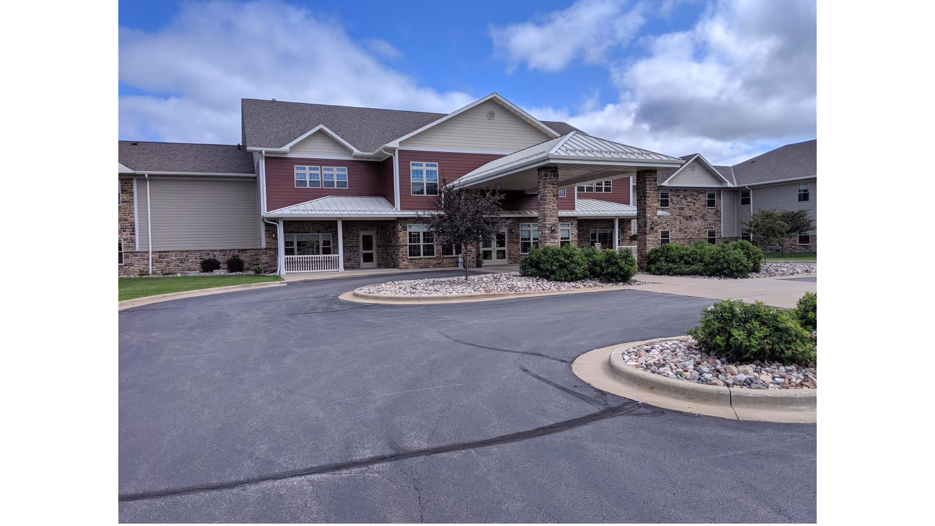 Two-story retirement community building with a covered entrance, circular driveway, and landscaped shrubs under a partly cloudy sky.