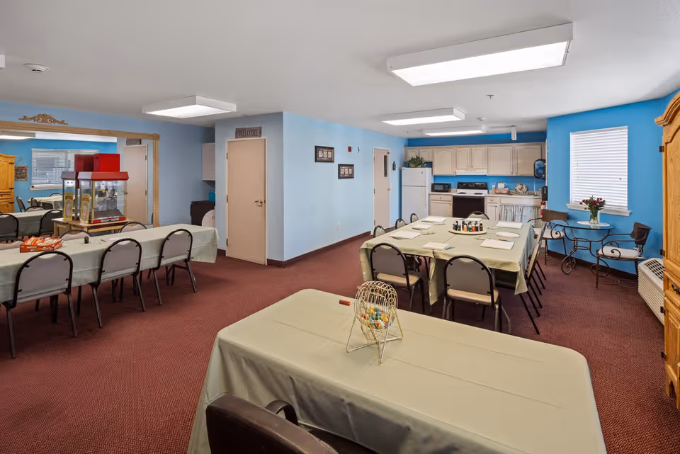 A senior living community common area with multiple tables covered in beige tablecloths and chairs arranged around them. The room has blue walls and red carpet. In the background, there is a kitchenette with white cabinets, a refrigerator, stove, and microwave. A popcorn machine is visible on one table, and a small round table with two chairs and a vase of flowers is near a window with blinds. The room is well-lit with ceiling lights.