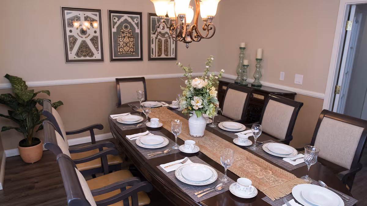 A formal dining room with a dark wooden table set for six people with white plates, cups, and wine glasses. A beige and brown table runner with a floral centerpiece in a white vase is in the middle of the table. The room has beige walls with white trim, three framed decorative artworks on the wall, a chandelier hanging above the table, a potted plant in the corner, and a sideboard with candles on it.