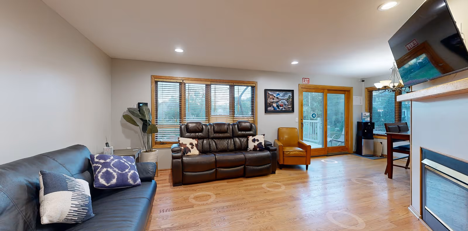 A cozy living room with wooden flooring, featuring a black leather sofa with two patterned pillows, a dark brown leather recliner sofa with two cowhide-patterned pillows, and a tan armchair. The room has large windows with wooden blinds, a glass door leading outside, a mounted flat-screen TV above a fireplace, a water dispenser, and a dining area with a table and chairs in the background.