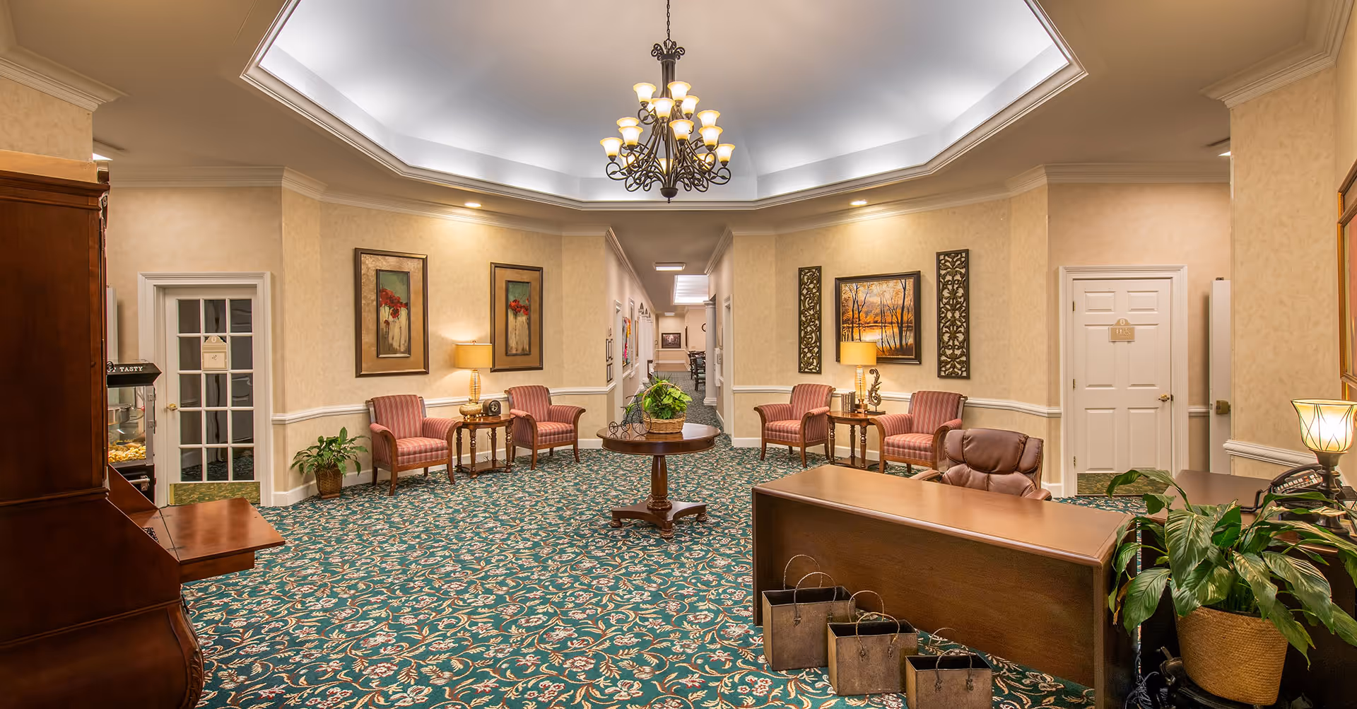 A warmly lit interior lobby area with patterned green carpet, beige walls, and a decorative chandelier hanging from a tray ceiling. The room features several upholstered chairs with side tables and lamps, framed artwork on the walls, a wooden reception desk with a leather chair, and a round table with a plant in the center. There are doors and a hallway visible in the background.