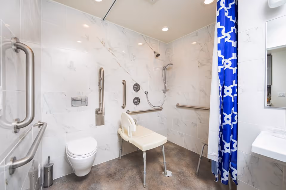 Accessible bathroom with white marble tiled walls, a wall-mounted toilet, multiple stainless steel grab bars, a shower area with a handheld showerhead, and a white shower chair. A blue and white patterned shower curtain is partially visible on the right side, along with a white sink and a mirror.