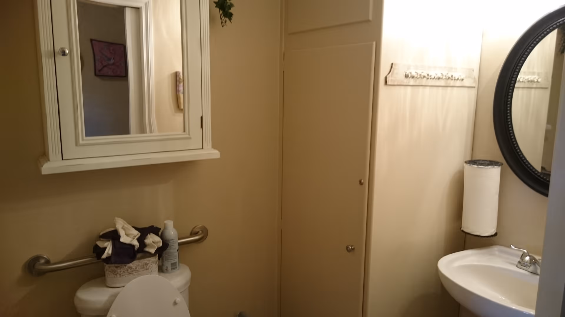 A small bathroom with beige walls featuring a white toilet with a grab bar behind it, a white wall-mounted cabinet with a mirror above the toilet, a round black-framed mirror above a white sink with a silver faucet, a paper towel holder with a roll of paper towels, and a wall-mounted rack with hooks.