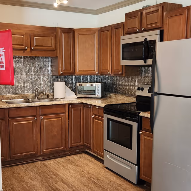 Small kitchen with brown wooden cabinets, stainless steel refrigerator, stove and microwave, a toaster oven on granite counters, and a tin-tile backsplash.