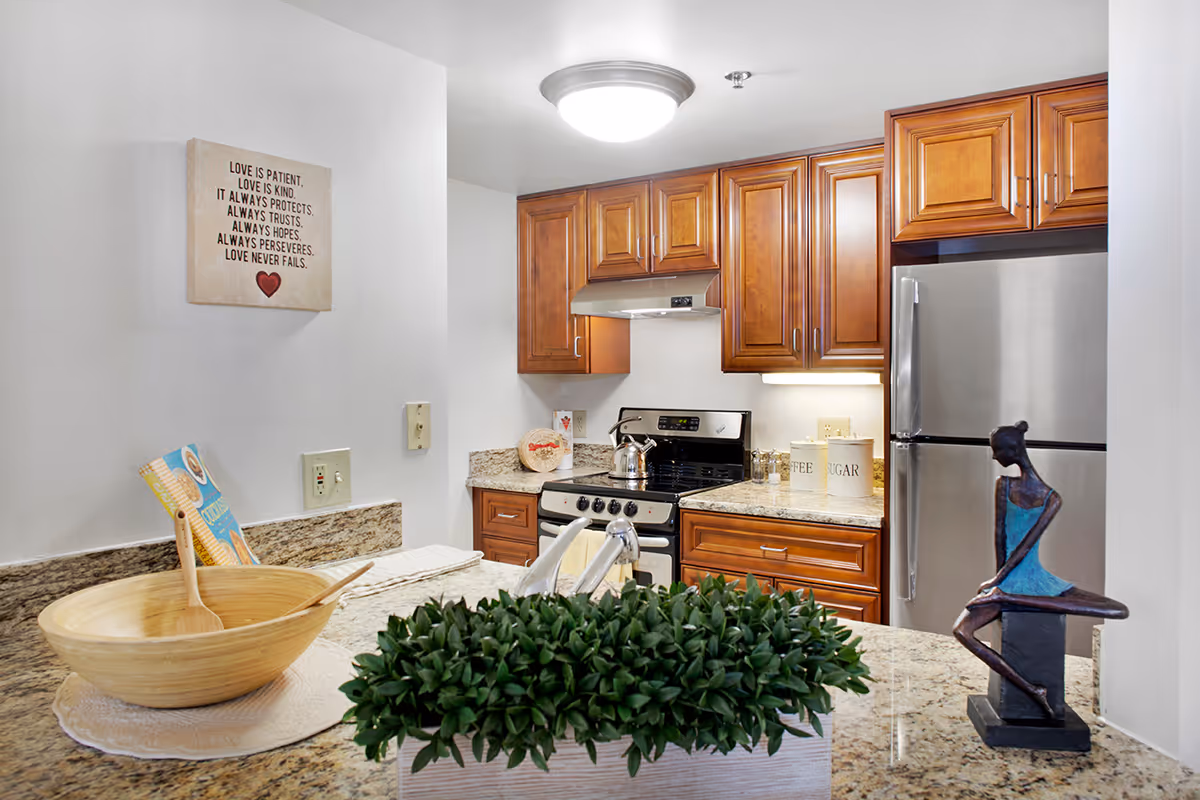 A modern kitchen with wooden cabinets, a stainless steel refrigerator, and a stove with a kettle on it. The countertop has a decorative green plant, a wooden bowl with a spoon, and a small sculpture of a seated figure. A wall decoration with an inspirational quote about love is visible on the left wall.