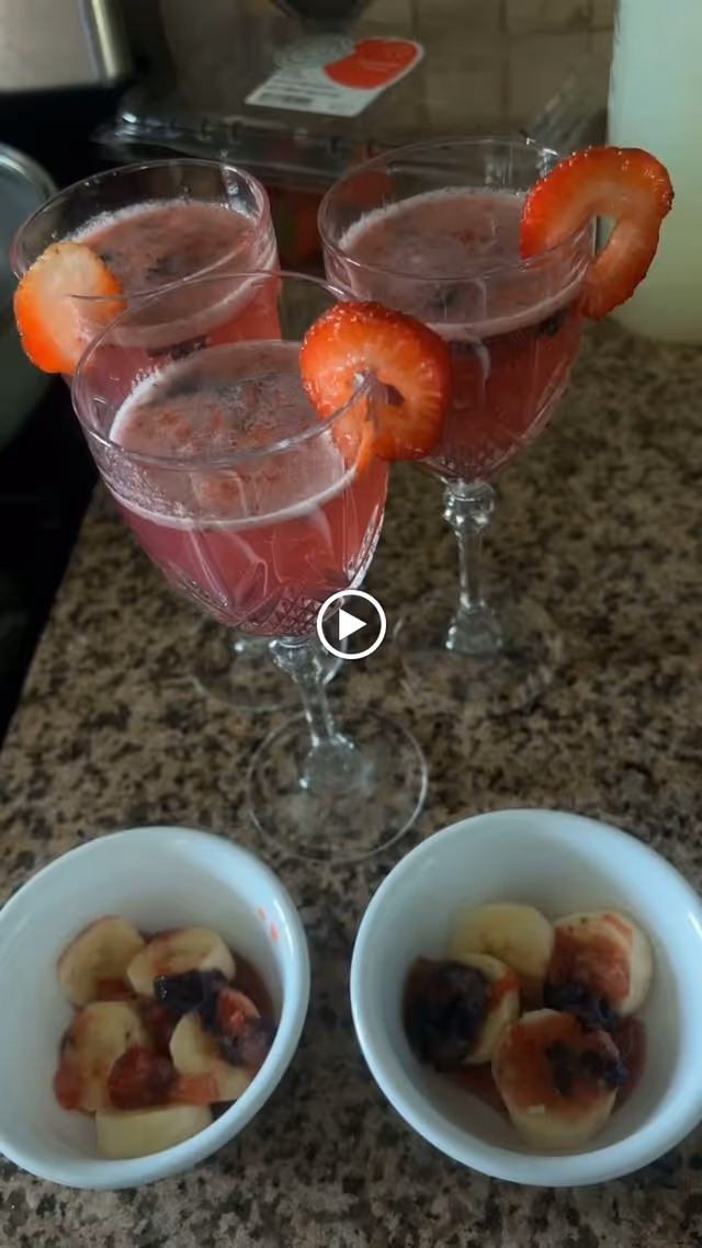 Three glasses filled with a pink beverage, each garnished with a slice of strawberry on the rim, placed on a granite countertop. In front of the glasses are two small white bowls containing sliced bananas topped with a dark red sauce or jam.