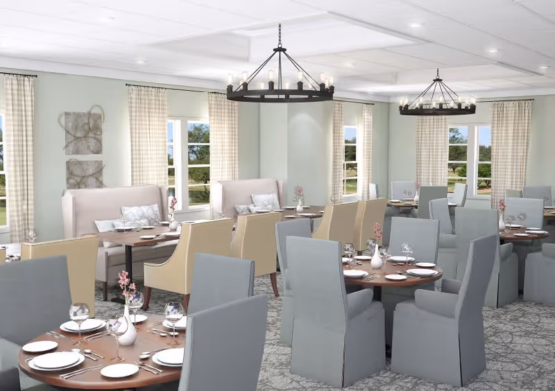 A bright and elegant dining room with multiple round and rectangular tables set with white plates, silverware, wine glasses, and small floral centerpieces. The room features light-colored walls, large windows with checkered curtains, cushioned chairs in beige and gray, and two black chandeliers hanging from the ceiling.