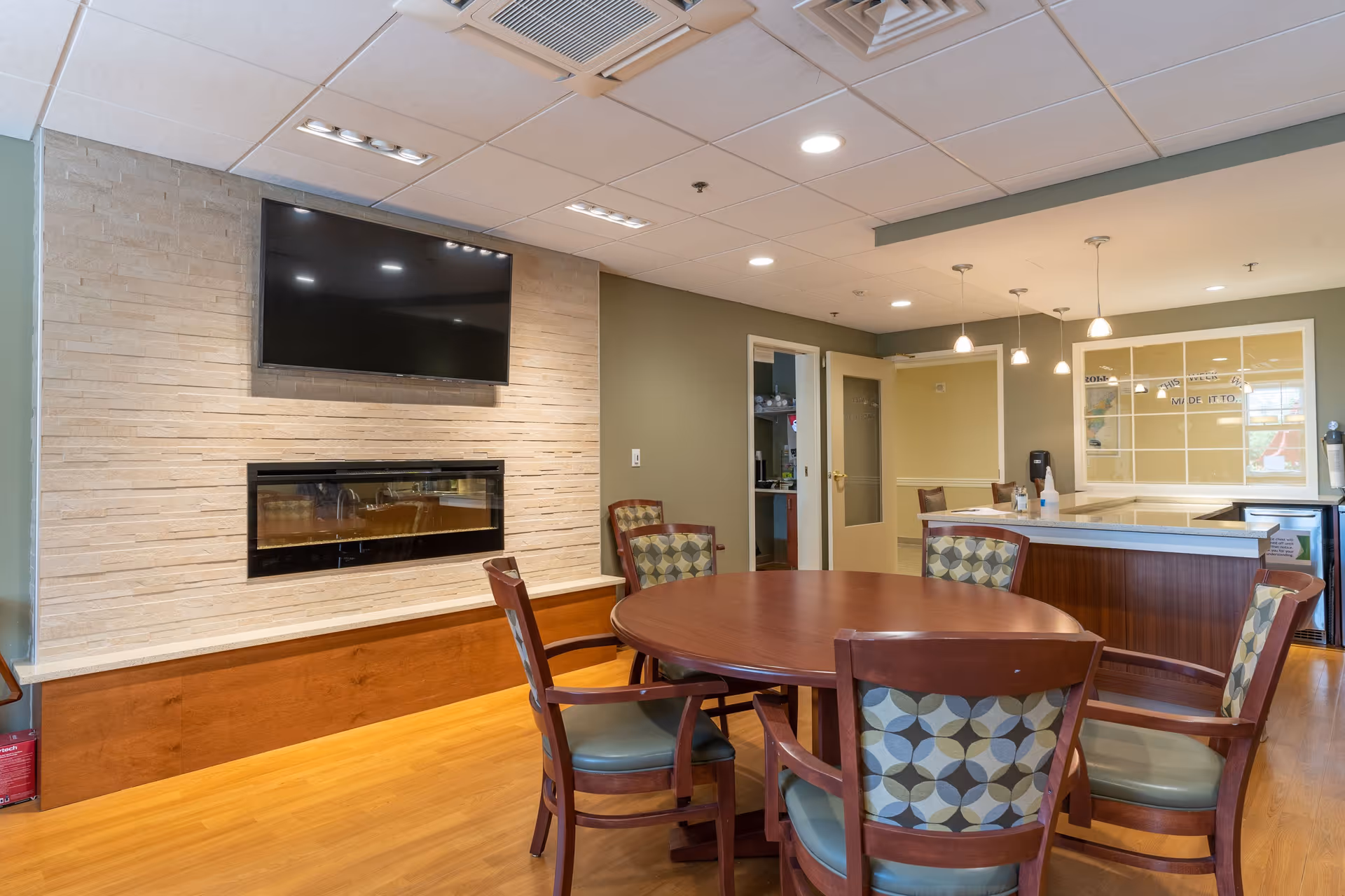 A cozy interior room with a round wooden table surrounded by six cushioned chairs with patterned backs. The room features a modern electric fireplace set into a stone accent wall beneath a mounted flat-screen TV. The floor is wooden, and the ceiling has recessed lighting and pendant lights hanging over a counter area in the background. There is an open doorway leading to another room and a window with a view into another interior space.