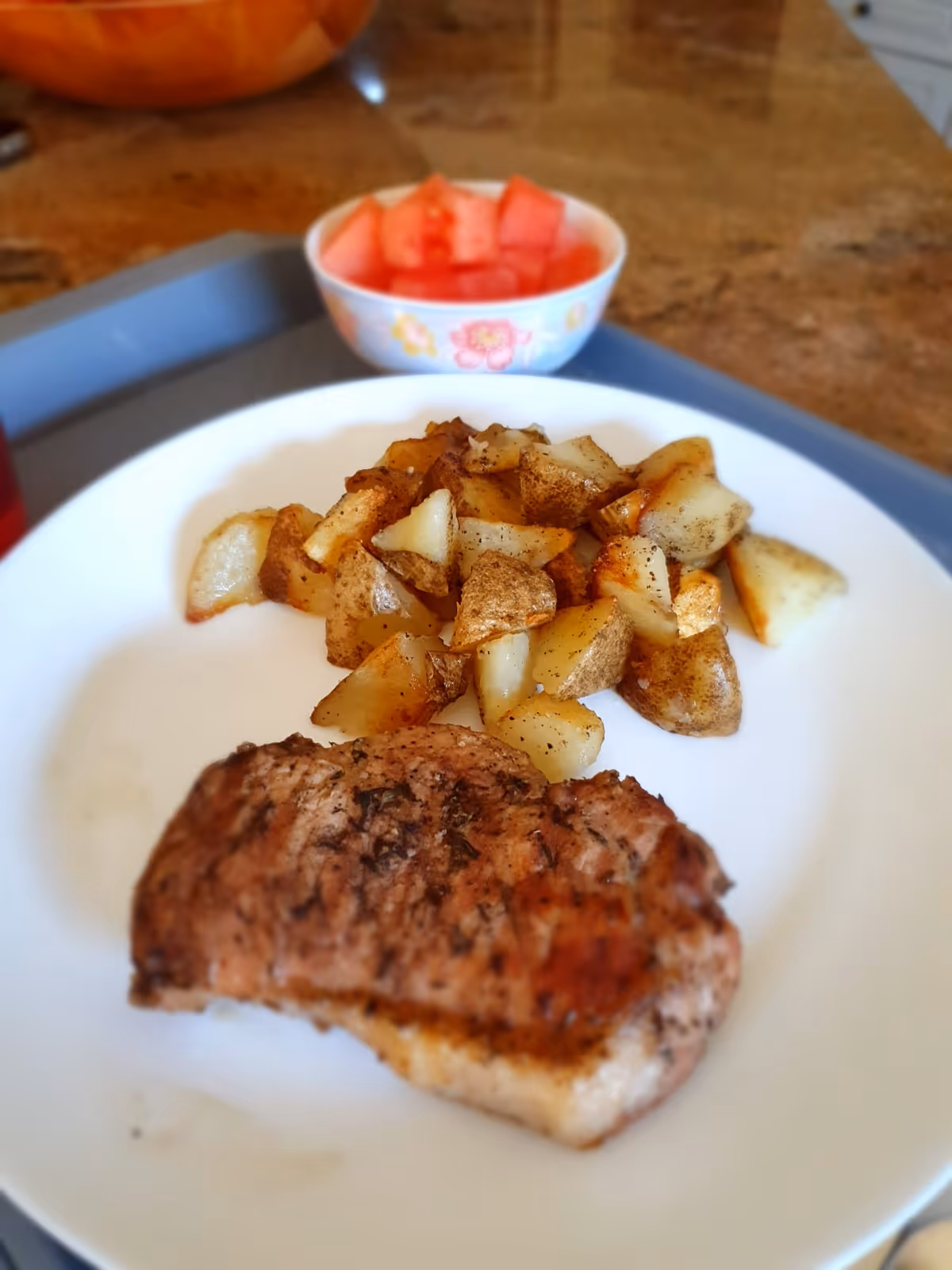 Plate with cooked meat and roasted potatoes on a tray, with a small bowl of watermelon in the background.