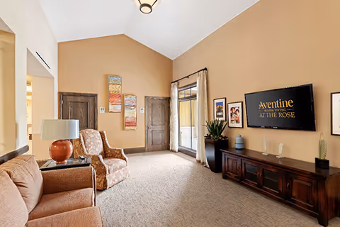 A cozy living room with beige walls and carpeted floor. The room features a brown sofa, a patterned armchair, a wooden side table with a lamp, and a dark wooden TV stand with a flat-screen TV mounted above it. The TV screen displays the text 'Aventine Memory Care at The Rose.' There are framed pictures on the wall and a large window with beige curtains letting in natural light.