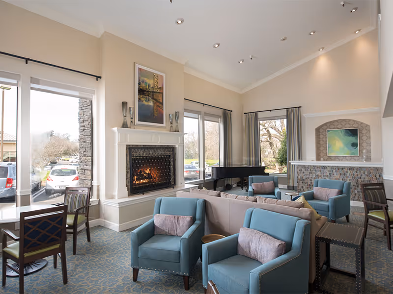 A bright and spacious living room area with large windows letting in natural light. The room features a lit fireplace with decorative vases on the mantel, a grand piano near the windows, and several comfortable blue armchairs and a beige sofa arranged around a small table. There are also a few dining chairs and tables near the windows, and a decorative tiled bar or counter area with artwork above it.