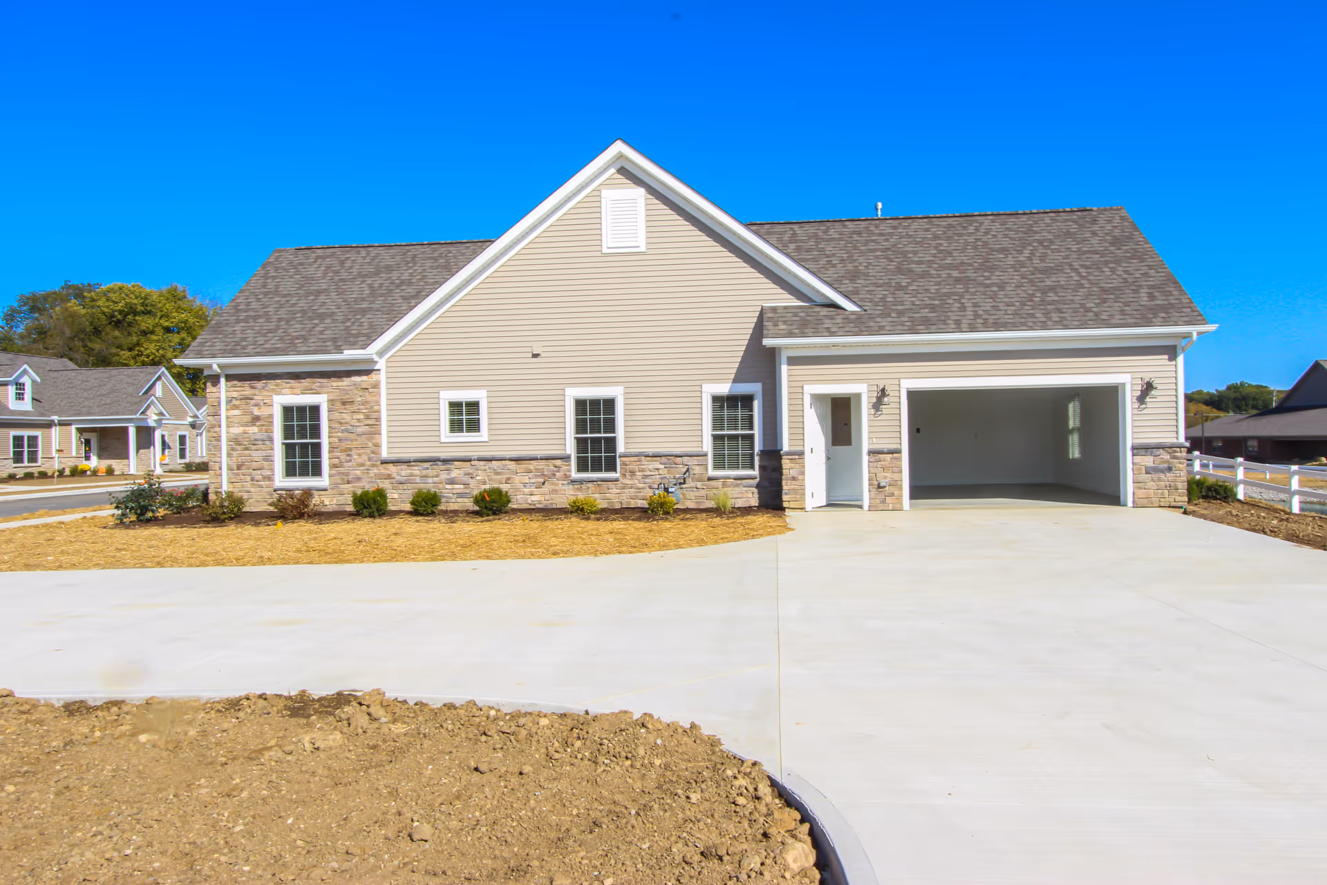 Single-story beige house with stone accents, an open two-car garage and a wide concrete driveway under a clear blue sky.