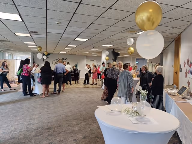 A group of people socializing in a large indoor room decorated with white and gold balloons. Several round tables are covered with white tablecloths and have floral centerpieces. The room has a carpeted floor and a drop ceiling with fluorescent lighting.