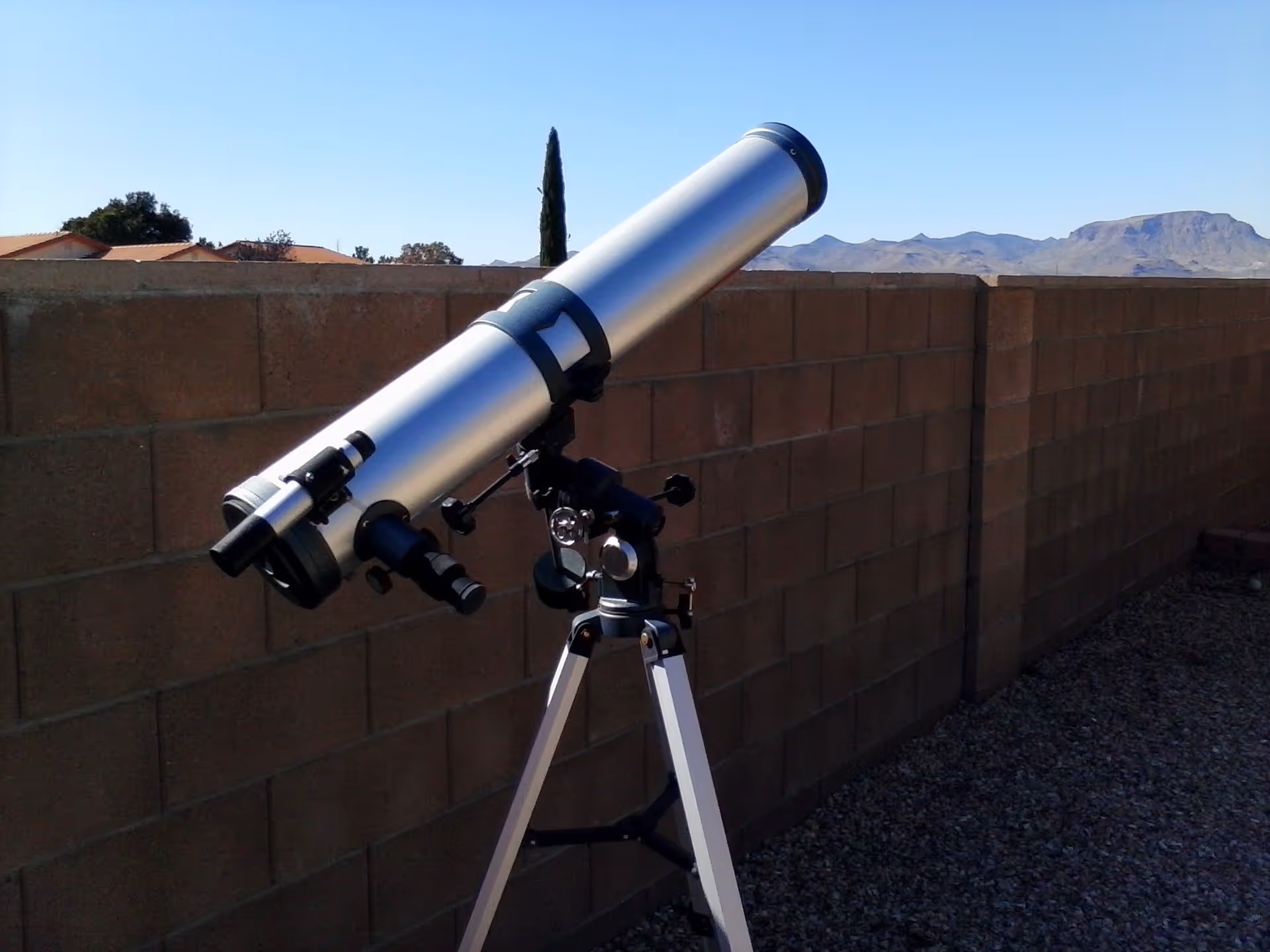 A silver telescope mounted on a tripod set up outdoors next to a brick wall with mountains visible in the background under a clear blue sky.