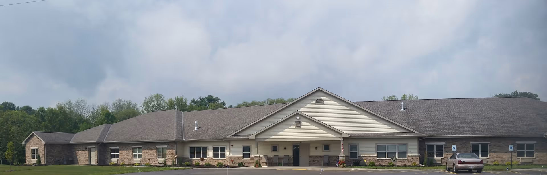Single-story building with a gable roof and brick and siding exterior, surrounded by green trees and a parking lot with a few cars, under a cloudy sky.