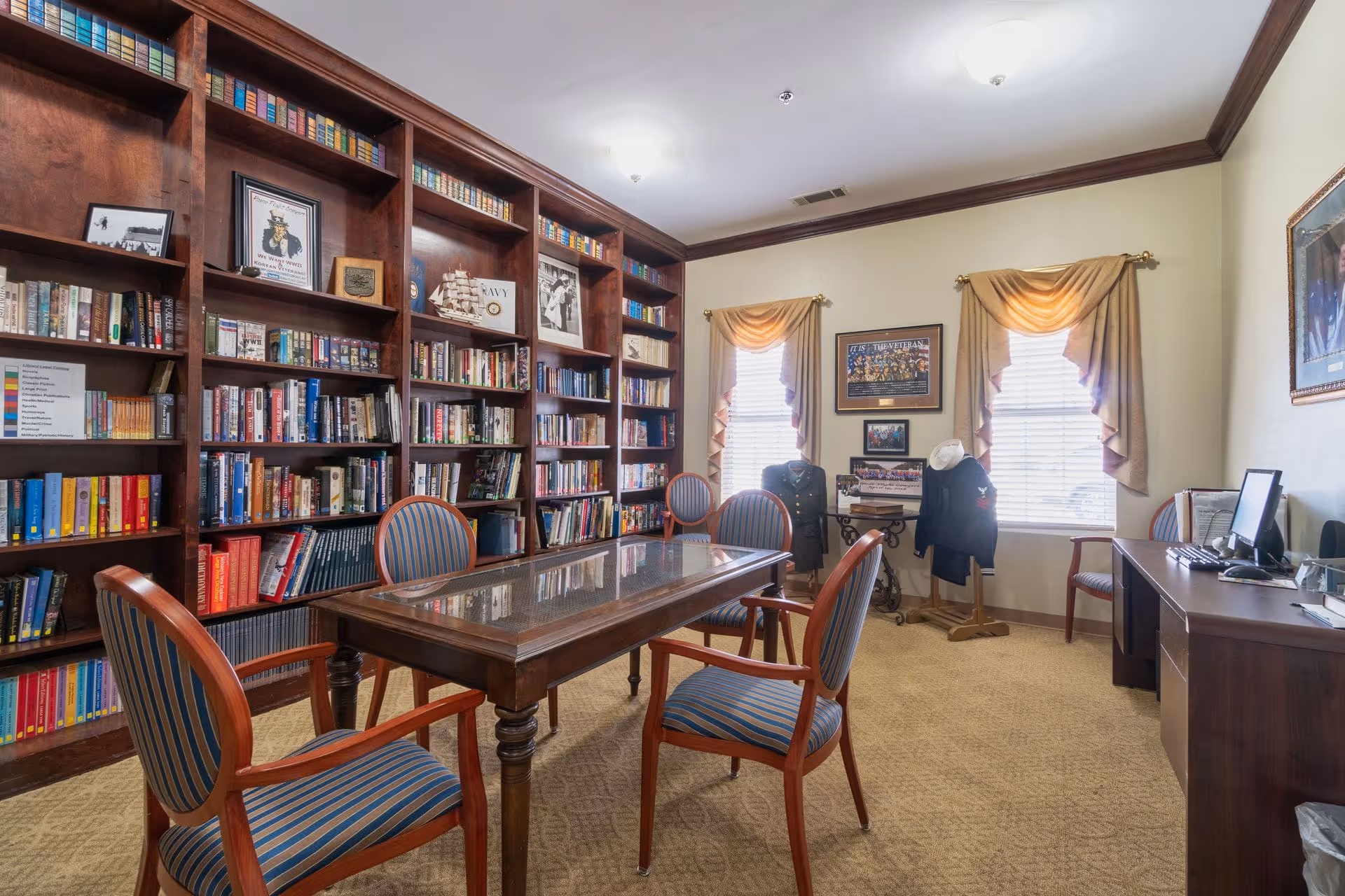 A cozy library room with wooden bookshelves filled with books and framed pictures. There is a wooden table with a glass top surrounded by four striped upholstered chairs. Two windows with beige curtains allow natural light into the room. A desk with a computer is positioned against the right wall, and military uniforms and memorabilia are displayed near the windows.