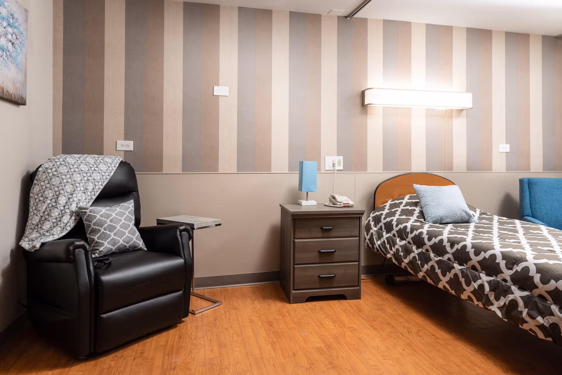 A cozy bedroom in a senior living facility featuring a single bed with patterned bedding and a blue pillow, a wooden nightstand with a blue lamp and a telephone, a black recliner chair with a patterned pillow and blanket, and striped wallpaper in shades of beige and brown.