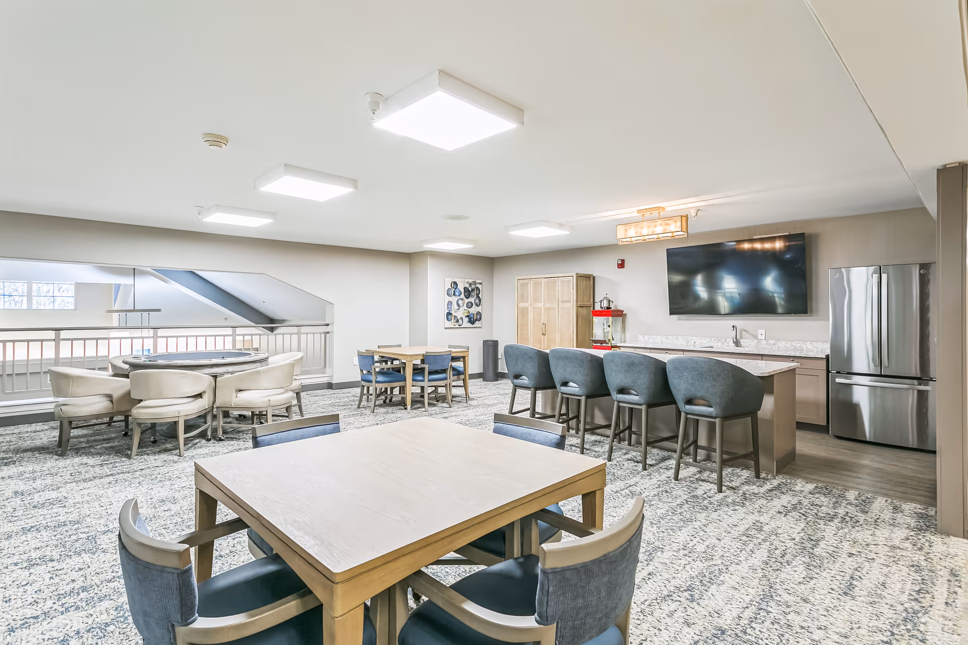 Community lounge and kitchenette with tables, bar seating, a wall-mounted TV, and a refrigerator in a senior living facility.