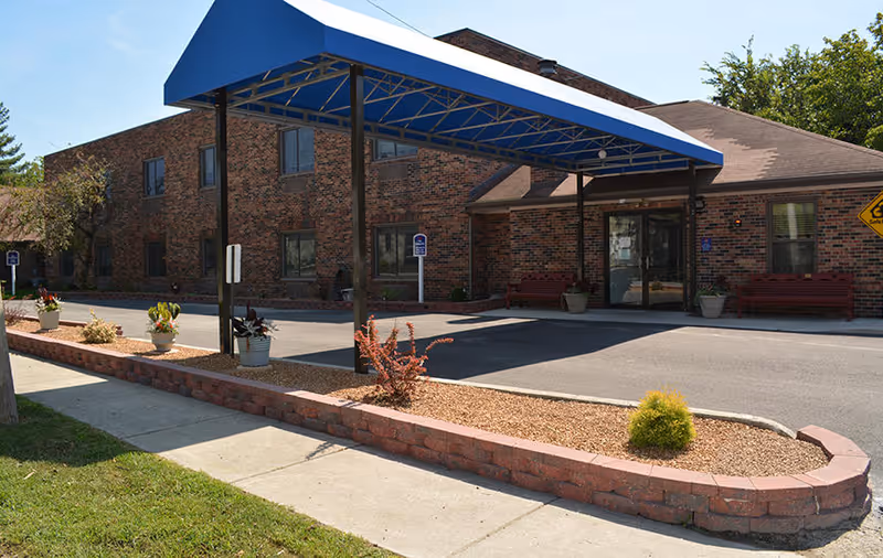 Exterior view of a brick building with a blue canopy over the entrance driveway. There are potted plants along a low brick wall and benches near the entrance. The scene is well-lit with clear skies and some trees in the background.