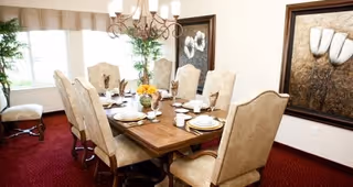 A formal dining room with a wooden dining table set for eight people. The chairs are upholstered in a light beige fabric. The room has a red carpet, large windows with beige curtains, a chandelier overhead, and two framed floral paintings on the walls.