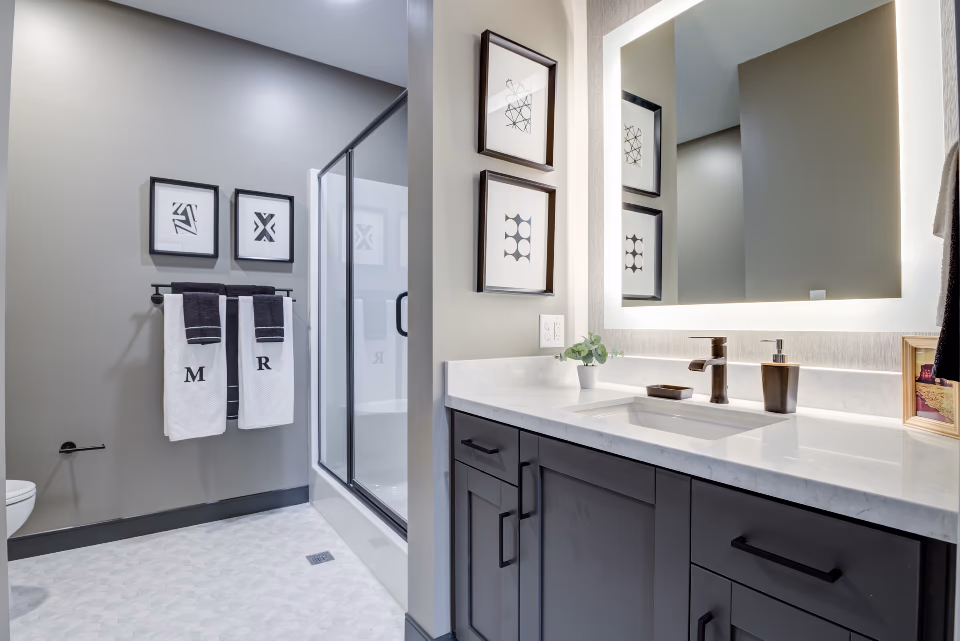 Modern bathroom with a glass-enclosed shower, two framed abstract art pieces on the wall, two white towels with initials M and R hanging on a towel rack, a large illuminated mirror above a white countertop with a sink, bronze faucet, soap dispenser, and a small potted plant. The cabinetry below the sink is dark gray.