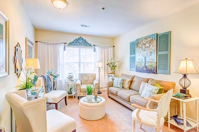 A bright and cozy living room with beige walls and carpet. The room features a beige sofa with multiple decorative pillows, a patterned armchair, two cream-colored chairs, and a round ottoman coffee table with a small plant on top. There are two side tables with lamps and decorative items, a large window with white curtains, and a large painting of a tree on the wall above the sofa.