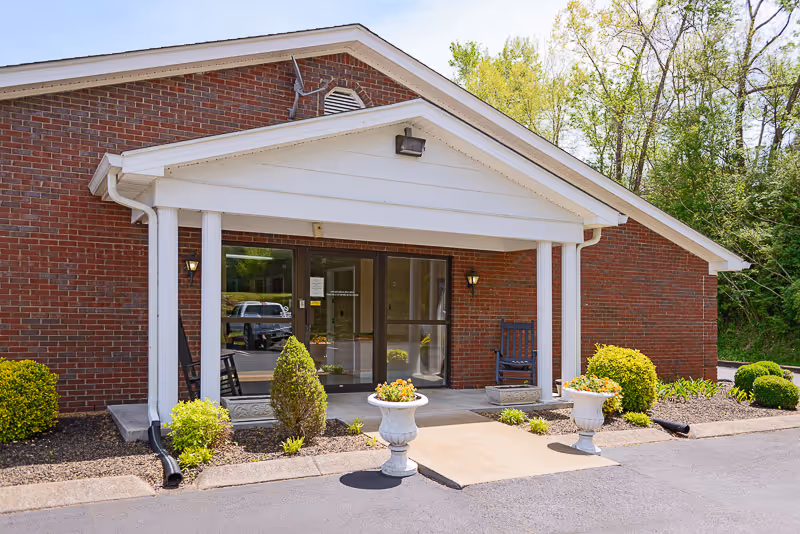 Entrance of a brick building with a white portico supported by four white columns. There are two black rocking chairs on the porch, two white planters with flowers, and neatly trimmed bushes along the walkway. Trees and greenery are visible in the background.
