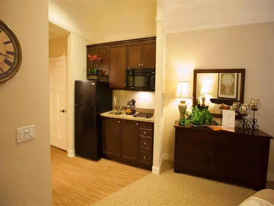 Small apartment-style kitchenette with dark cabinets, a black refrigerator and microwave, and an adjacent dresser with a lamp and mirror.
